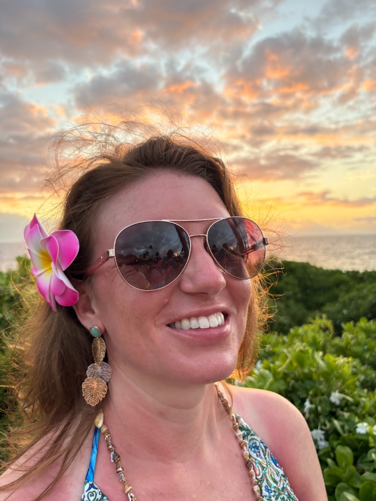 Sarah, the founder of Totes of Love, smiling warmly with a pink flower in her hair and a sunset over the ocean behind her