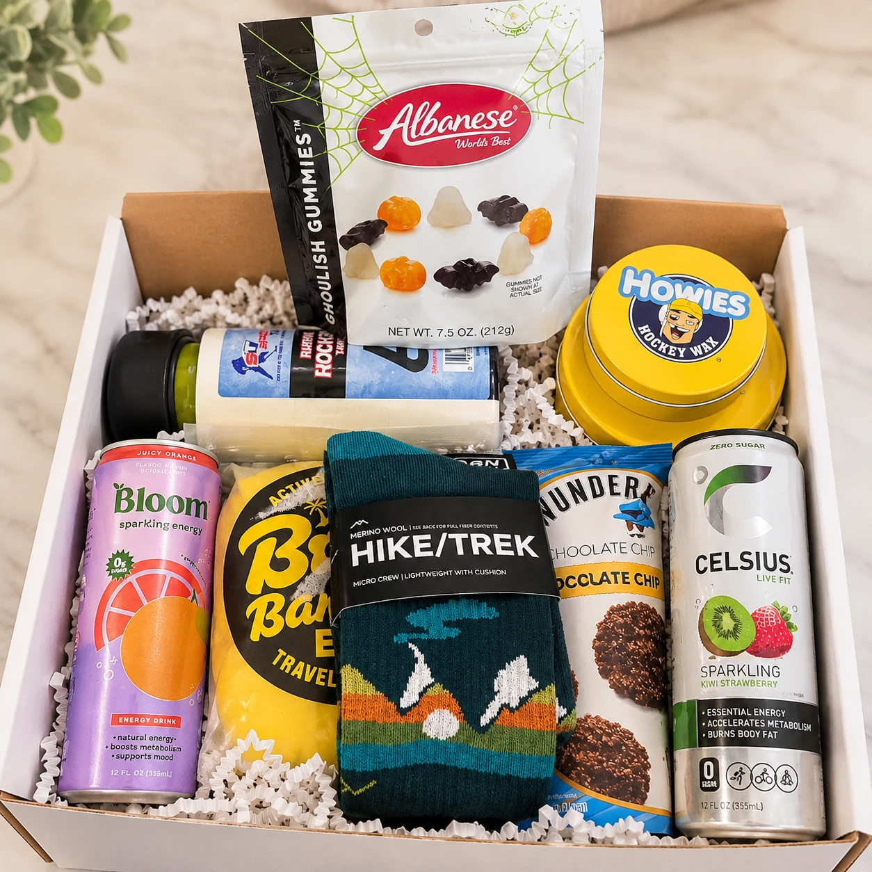 An open white gift box packed for a student who plays hockey and hikes: hockey stick wax and tape, merino hiking socks, sparkling energy drinks, chocolate chip cookies and a bag of gummies, on a marble counter.